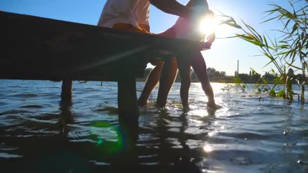 Padre che tiene tra le mani il figlio mentre siede sul bordo del molo di legno al lago e penzola le gambe in acqua. Il bambino e suo padre dondolavano i piedi nel fiume nella giornata di sole. Rallentatore Close up — Video Stock