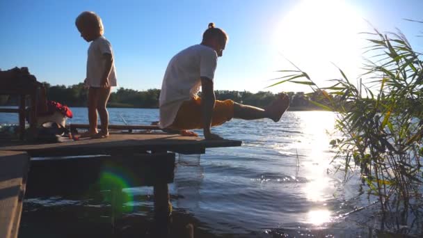 Молодой человек практикует йогу на краю деревянного причала у озера в солнечный летний день. Маленький ребенок и его отец проводят время вместе на природе. Здоровый активный образ жизни. Медленное движение — стоковое видео