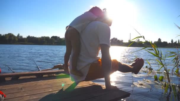 Ein junger Mann praktiziert Yoga mit seinem Sohn auf dem Rücken am Rande eines hölzernen Stegs am See. Die Familie verbringt an einem Sommertag gemeinsam Zeit in der Natur. gesunder aktiver Lebensstil. Zeitlupe aus nächster Nähe — Stockvideo