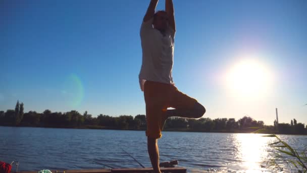 Joven practicando yoga en el borde del embarcadero de madera en el lago en el día de verano. Hombre deportivo haciendo ejercicio cerca del lago con luz solar en el fondo. Vida activa saludable. Cámara lenta Primer plano — Vídeo de stock