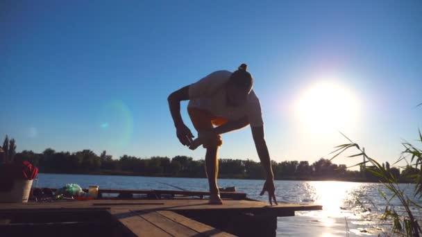 Giovane ragazzo che pratica mosse yoga e posizioni sul bordo del molo di legno al lago nella giornata di sole. Uomo sportivo che si allena a contatto con la natura con la luce solare sullo sfondo. Stile di vita sano e attivo. Rallentatore Close up — Video Stock