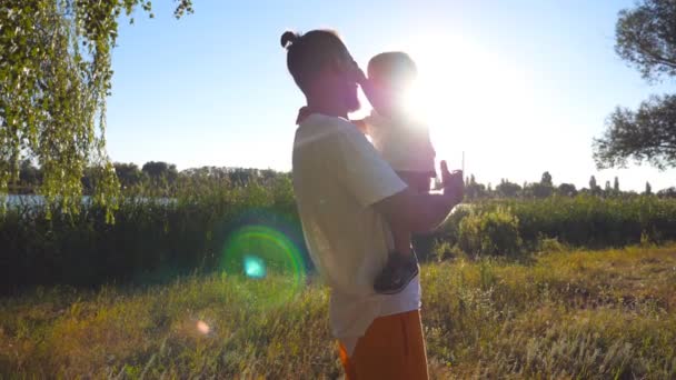 Heureux papa tenant dans ses mains son fils aux cheveux blonds au parc par une journée ensoleillée. Le jeune père et son petit enfant passent du temps ensemble et apprécient la nature estivale. Vue latérale Mouvement lent Fermer — Video