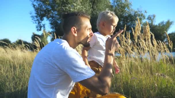 日当たりの良い夏の日に草でブロンドの髪とかわいい息子と座っている若いお父さんのプロフィール。幸せな家族が自然で一緒に時間を過ごします。背景の美しい自然。スローモーションをクローズ アップ — ストック動画