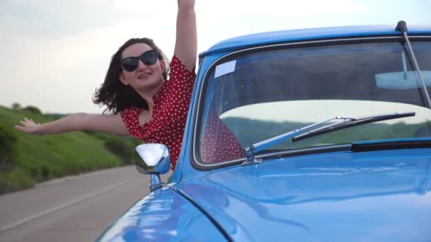 Menina sorridente jovem em óculos de sol inclinando-se para fora da janela do carro vintage e desfrutar de viagem. Mulher atraente feliz olha para fora de mover auto retro e levantar as mãos. Conceito de viagem e liberdade. Movimento lento — Vídeo de Stock