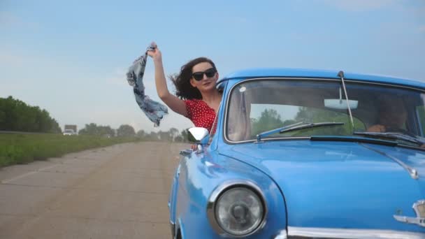 Young girl with scarf in hand leaning out of vintage car window and enjoying trip. Woman looks out of moving retro car with beautiful landscape at background. Travel concept. Slow motion Close up — Stock Video