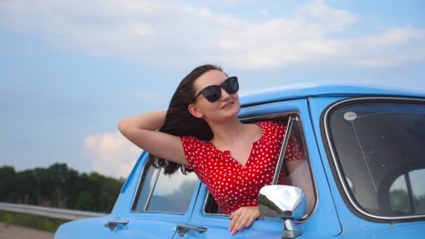 Chica joven en gafas de sol que se inclina por la ventana del coche vintage y disfrutar del viaje. Mujer mira hacia fuera de mover auto retro. Concepto de viaje y libertad. Fondo borroso. Movimiento lento — Vídeos de Stock