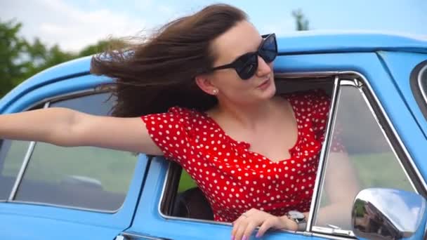 Ung flicka i solglasögon lutar ut vintage bilfönstret och njuta av resan. Kvinnan ser från rörliga retro auto. Resor-konceptet. Suddig bakgrund. Slow motion närbild — Stockvideo