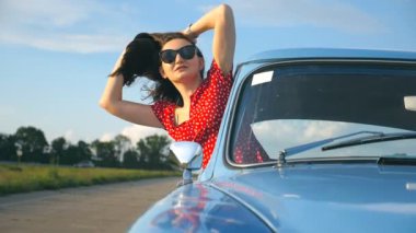 Genç kız eski model araba penceresinden yaslanmış ve yolculuk zevk güneş gözlüğü. Kadın retro otomatik hareket etmesini bakıyor. Seyahat ve Özgürlük kavramı. Arka plan bulanık. Önden görünüm ağır çekim