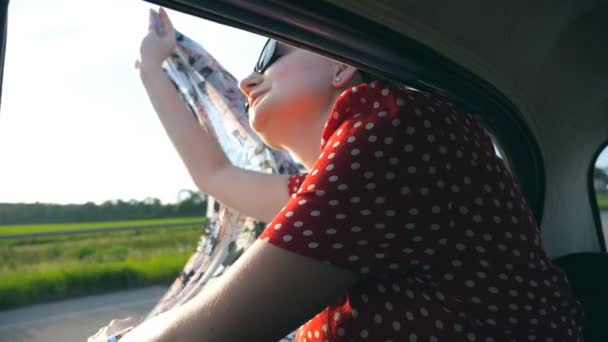 Joven chica atractiva en gafas de sol con bufanda en las manos apoyada en el coche de la vendimia ventana y disfrutando de paseo en verano. Concepto de viaje y libertad. Vista interior cámara lenta Primer plano — Vídeos de Stock