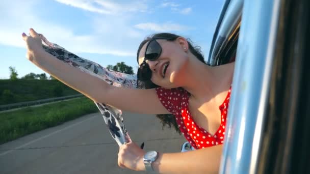 Chica joven en gafas de sol con bufanda en las manos que se inclina por la ventana coche retro y el viaje de regocijo. Mujer atractiva sonríe y disfruta de viaje en día soleado. Concepto de viaje. Cámara lenta Primer plano — Vídeos de Stock