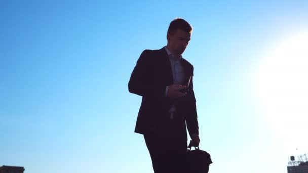 Joven hombre de negocios con un maletín esconde el teléfono en el bolsillo del traje y caminar en la ciudad con el cielo azul en el fondo. Un hombre de negocios viajando al trabajo. Un tipo seguro que está de camino al trabajo. Movimiento lento — Vídeo de stock