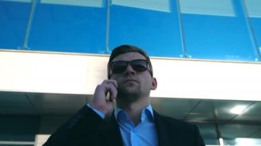 Güneş gözlüklü genç işadamının portresi ofisin dışında telefonla konuşuyor. Arka planda modern iş binasıyla cep telefonuyla konuşan yakışıklı iş adamına yakın çekim. Yavaş çekim.