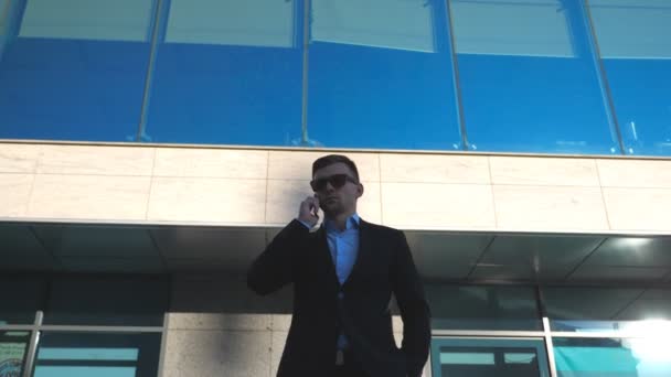 Porträtt av ung affärsman i solglasögon talar på telefon utomhus nära kontoret. Närbild av stilig affärsman talar på mobiltelefon med moderna affärsbyggnad i bakgrunden. Långsamma rörelser — Stockvideo
