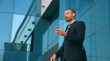 Genç işadamı ofis yakınında akıllı telefon kullanıyor ve başarıyı kutluyor. İş adamı telefonda iyi haberleri ve olumlu duyguları öğrendi. Erkek portresi başarıyla mutludur. Yavaş çekim