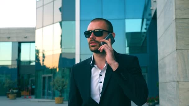 Porträtt av affärsman i solglasögon som pratar i telefon och går på gatan. Ung man som pratar affärer under resan till jobbet. En självsäker kille i kostym på väg till kontoret. — Stockvideo