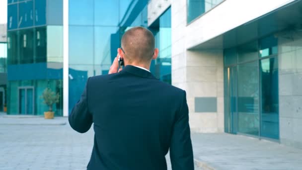 Jonge zakenman praten over telefoon en wandelen in de straat. Onherkenbaar man met zakelijke gesprek tijdens het woon-werkverkeer te werken. Zeker man in pak wordt op zijn weg naar kantoorgebouw. Achteraanzicht — Stockvideo