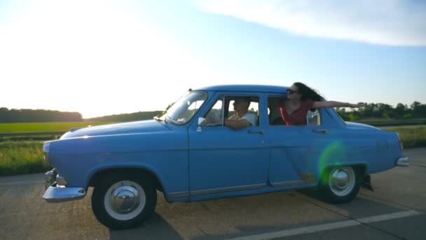 Ragazza attraente in occhiali da sole appoggiata al finestrino dell'auto d'epoca e godendo di viaggio. Giovani coppie viaggiano su auto retrò in estate. Concetto di viaggio. Sole che splende sullo sfondo. Rallentatore Close up — Video Stock