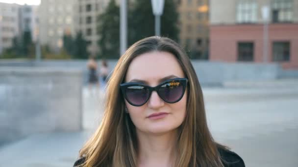 Retrato de una joven empresaria en gafas de sol caminando por la calle de la ciudad. Atractiva mujer de negocios mirando a la cámara. Rostro de chica confiada viajando al trabajo. Primer plano: cámara lenta — Vídeos de Stock