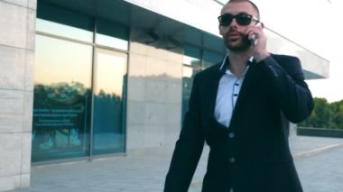 İşadamı caddede yürüyüş ve telefonda konuşurken güneş gözlüğü. Çalışmaya commuting sırasında Telefonda konuşan genç işadamı. Açık konuşmayı elbiseli yakışıklı adam. Ağır çekim