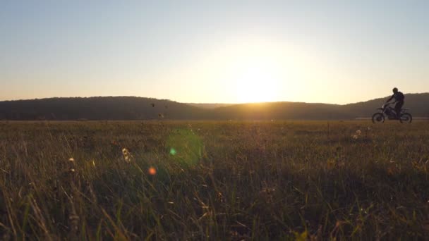 Motorradfahrer auf dem Weg durch ein großes Feld mit wunderschönem Sonnenuntergang im Hintergrund. Die warme Sommersonne erhellt die grüne Vegetation des Feldes. Konzept der aktiven Erholung von der Natur. Zeitlupentempo — Stockvideo