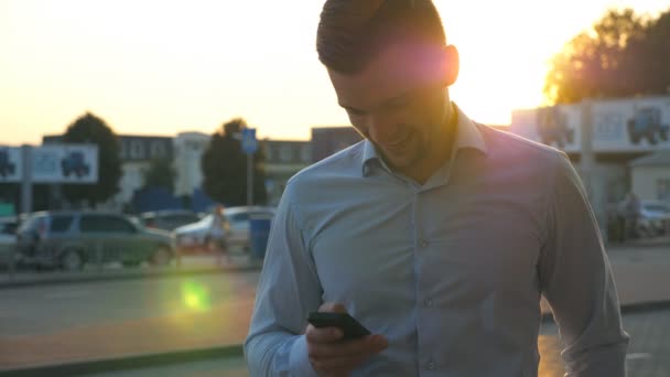 接近年轻成功的商人穿着衬衫站在城市环境中，日落时使用智能手机。一个英俊的男人在城市街道的手机上留言。背景下的阳光 — 图库视频影像