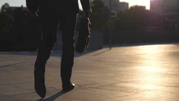 Pies de joven hombre de negocios con un maletín caminando por la calle de la ciudad. Un hombre de negocios viajando al trabajo. El tipo de confianza en traje va camino al trabajo. Fondo del paisaje urbano. Vista trasera en cámara lenta Primer plano — Vídeo de stock