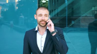 Ofisin yakınında telefonla konuşan yakışıklı iş adamının portresi. Genç iş adamı cep telefonuyla konuşuyor ve olumlu duygular besliyor. Adam sohbetten memnun. Yavaş çekim. Yakın çekim.