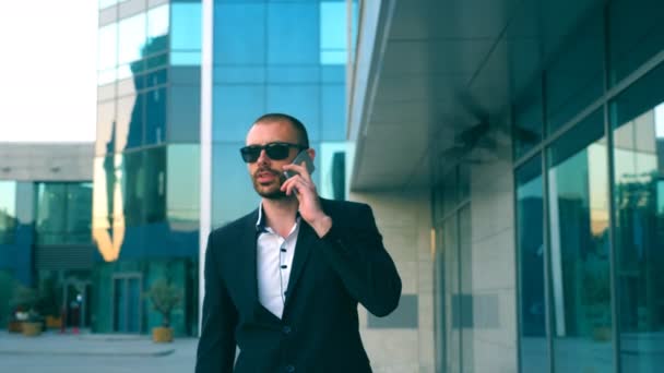 Retrato de hombre de negocios en gafas de sol hablando por teléfono y caminando por la calle. Joven hombre teniendo conversación de negocios durante el viaje al trabajo. Confiado en que el tipo del traje está de camino al edificio de oficinas — Vídeo de stock