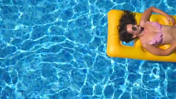 Vista superior da menina bronzeada bonita em óculos de sol e biquíni rosa deitado no colchão inflável amarelo na piscina. Jovem relaxante na bacia do hotel durante as viagens de verão. Conceito de férias — Vídeo de Stock
