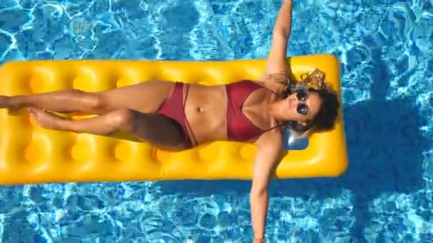 Девушка в солнцезащитных очках и красном бикини лежит на желтой надувной матрешке в бассейне. Молодая загорелая женщина отдыхает в бассейне отеля в солнечный день. Концепция отпуска. Вид сверху — стоковое видео