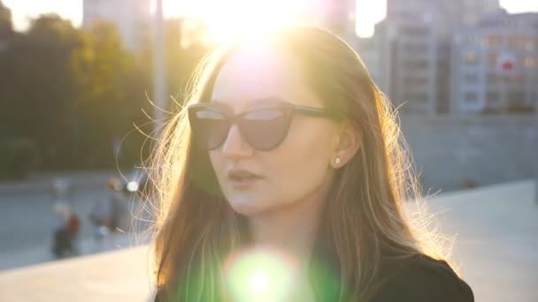 Портрет молодой предпринимательницы в солнечных очках, гуляющей по городской улице с солнечной вспышкой на заднем плане. Лицо привлекательной деловой женщины, приезжающей на работу. Закат освещает волосы девушки. Закрыть — стоковое видео