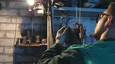 Gözlüklü erkek mekanik araç ayrıntı dikkatle inceliyor. Repairer garaj veya atölye içinde çalışır. Çalışan aracı işi için kullanır. Yan görünümü ağır çekim yakın çekim