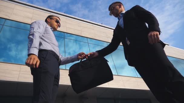 Un exitoso hombre de negocios pasando un maletín negro a su socio cerca del edificio de oficinas. Jóvenes colegas varones estrechando las manos en el entorno urbano. Negocios apretón de manos al aire libre. Vista de ángulo bajo Cámara lenta — Vídeo de stock