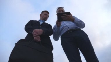 Genç iş adamı, şehirdeki meslektaşına tablet ekranında bir sunum gösteriyor. Başarılı girişimciler arka planda mavi gökyüzü olan dijital tablet üzerinde çalışıyorlar. Düşük açı görünümü Yavaş çekim