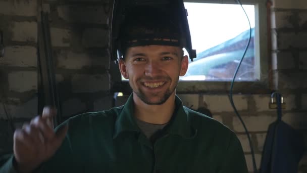 Saldatore sorridente guarda nella fotocamera e abbassa la maschera per mettersi al lavoro. Uomo felice con la barba che lavora nel suo garage o laboratorio. Il ragazzo ride e si gode la vita. Rallentatore Close up — Video Stock