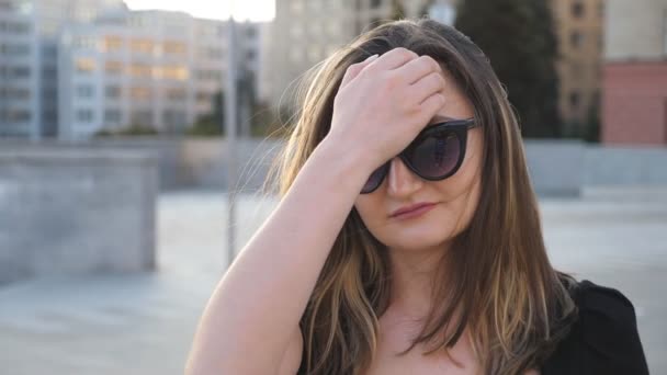 Portrait of young businesswoman in sunglasses walking in city street. Attractive business woman looking at camera and straightens hair. Face of confident girl commuting to work. Close up Slow motion — Stock Video