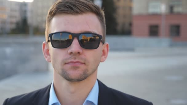 Retrato de un joven empresario con gafas de sol caminando por la calle de la ciudad. Hombre de negocios guapo mira la cámara paseando por la plaza de la ciudad. La cara de un tipo seguro que viaja al trabajo. Cámara lenta Primer plano — Vídeo de stock