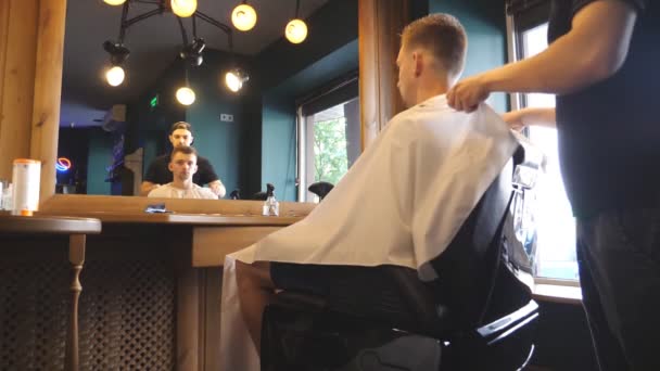 Male hairdresser finished to perform a haircut and taking off salon cape from his client at salon. Young barber removes protective cloak from his customer in barbershop. Reflection of men in mirror — Stock Video