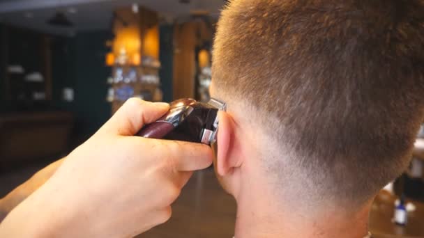 Bras de coiffeur faisant coupe masculine au client avec tondeuse dans le salon à la mode. Mains de coiffeur couper les cheveux du gars avec rasoir électrique dans le salon de coiffure. Processus de coiffure. Vue arrière Mouvement lent — Video