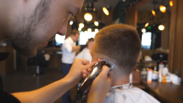 El peluquero profesional que corta los cabellos del muchacho con la maquinilla de afeitar eléctrica en la barbería. Peluquería haciendo corte de pelo masculino para el cliente con clipper en el salón. El jefe de cliente está borroso. Primer plano: cámara lenta — Vídeo de stock