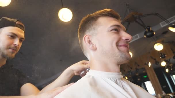 Перукар використовує щітку для чищення клієнта нарізаного волосся в перукарні. Чоловічий перукар використовує тальк на шиї молодого хлопця в салоні. Перукар робить косметичні процедури для щасливого клієнта. Повільний рух — стокове відео