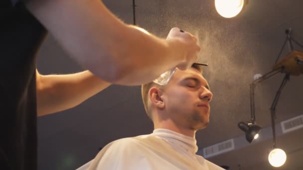 Gros plan du jeune coiffeur pulvérisant de l'eau sur les cheveux masculins du pulvérisateur après la coupe dans le salon. Coiffeur peigner les cheveux de son client dans le salon de coiffure. Processus de coiffure. Vue latérale Mouvement lent — Video