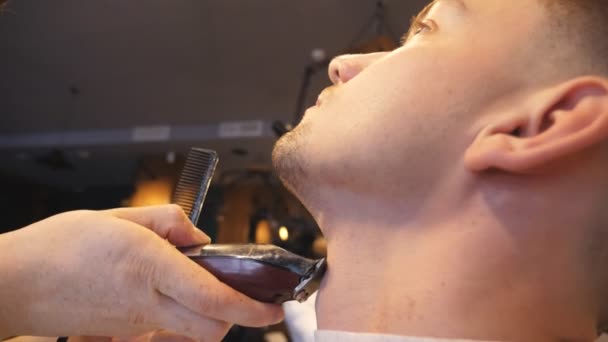 Férfi kezében professzionális fodrász tisztítás szakáll fiatal vevő elektromos borotva, a fodrászatban. Fodrász borotválkozás ügyfele Trimmer címere. Fodrász folyamat. Oldalnézet lassú mozgás — Stock videók