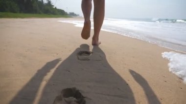 Kadın ayaklar arka planda okyanus dalgaları ile plajda altın kum üzerinde yürüyor. Genç kadının bacakları kuma basıyor. Deniz kıyısında yalınayak kız. Yaz tatili tatili. Yavaş hareket yakın çekim