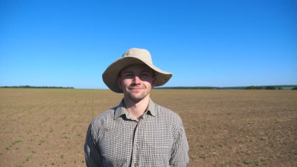 Retrato de joven granjero feliz con sombrero mirando a la cámara y sonriendo sobre el fondo del campo y el cielo azul. Primer plano del hombre guapo con camisa de pie en el prado en un día soleado de verano — Vídeos de Stock