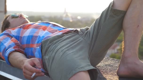 Handsome man in sunglasses lying on roof and holding cigarette in his hand with cityscape at background. Young guy relaxing and blowing smoke outdoor. Beautiful view Dolly shot Slow motion Close up — Stock Video