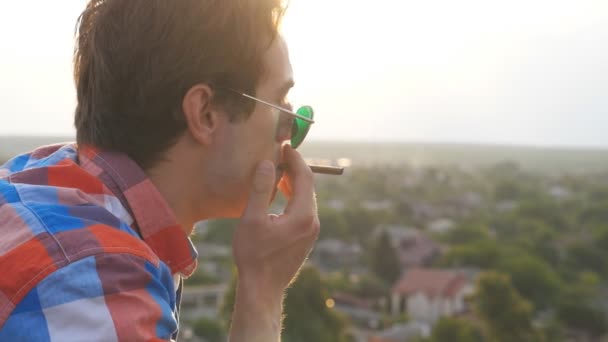 Profil eines gutaussehenden Mannes mit Sonnenbrille, der auf dem Dach steht und Zigarette raucht, mit Stadtbild im Hintergrund. junger Kerl, der sich ausruht und den Moment im Freien genießt. Schöne Aussicht. Zeitlupe aus nächster Nähe — Stockvideo