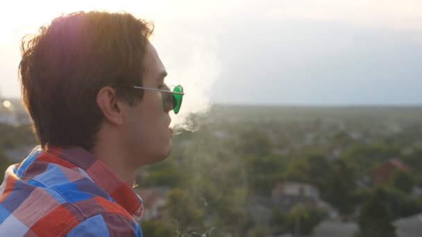 Perfil del hombre guapo en gafas de sol de pie en el techo y soplando humo en el fondo del paisaje urbano. Joven descansando y disfrutando del momento por la noche. Hermosa vista. Cámara lenta Primer plano — Vídeos de Stock