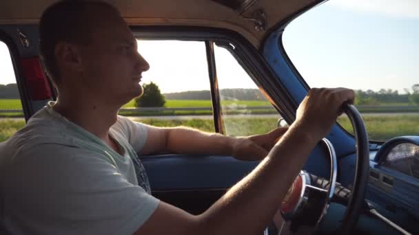 Profilo del giovane che si tiene per mano sul volante e guida un'auto retrò con brillamento del sole sullo sfondo. Un ragazzo caucasico che viaggia in un veicolo d'epoca in campagna. Vista laterale Rallentatore Close up — Video Stock