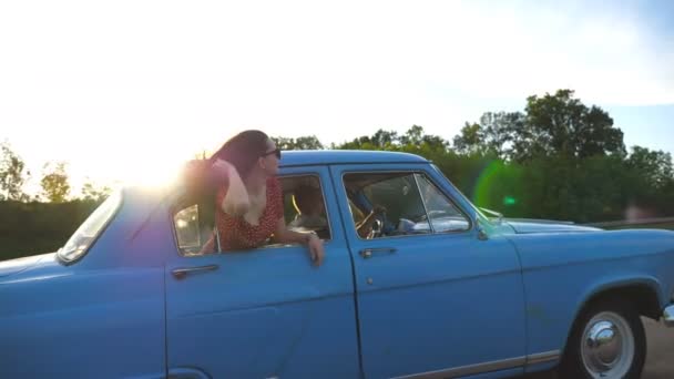 Chica feliz en gafas de sol que se inclina por la ventana del coche retro y disfrutar del viaje. Mujer joven mirando por la ventana de mover viejo auto. Resplandor solar en segundo plano Concepto de viaje y libertad. Cámara lenta Primer plano — Vídeo de stock